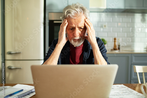 Worried senior man checking bills on laptop at kitchen table