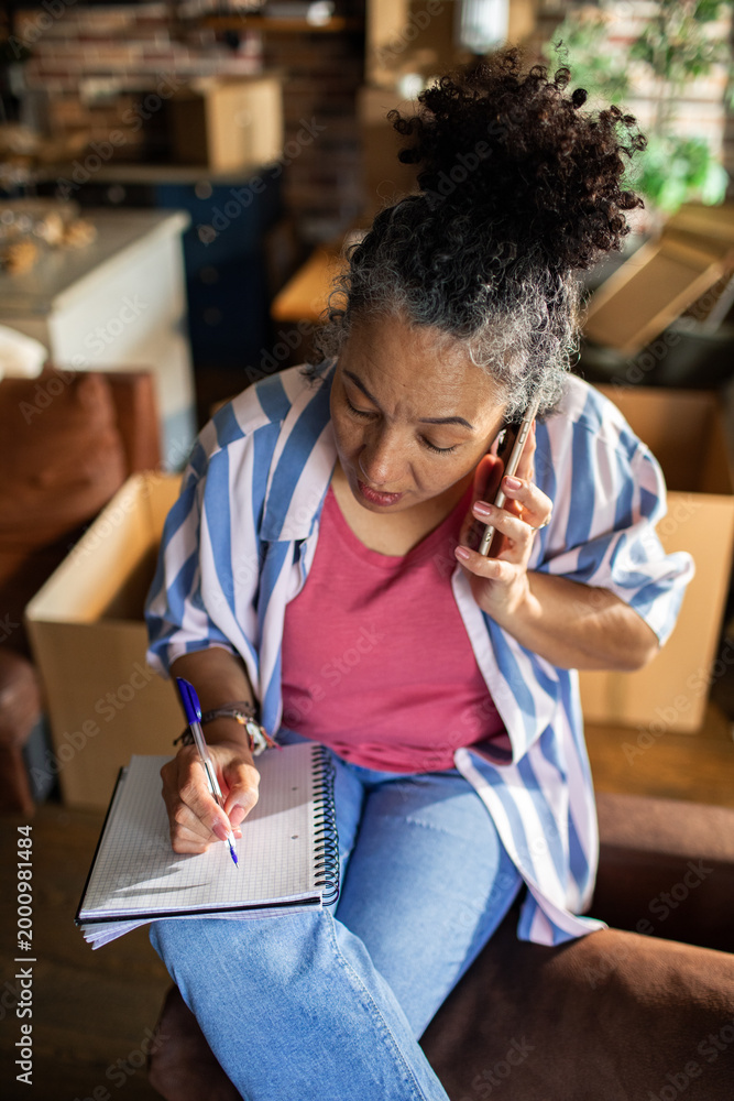 Naklejka premium Middle-aged woman on phone taking notes while planning move at home