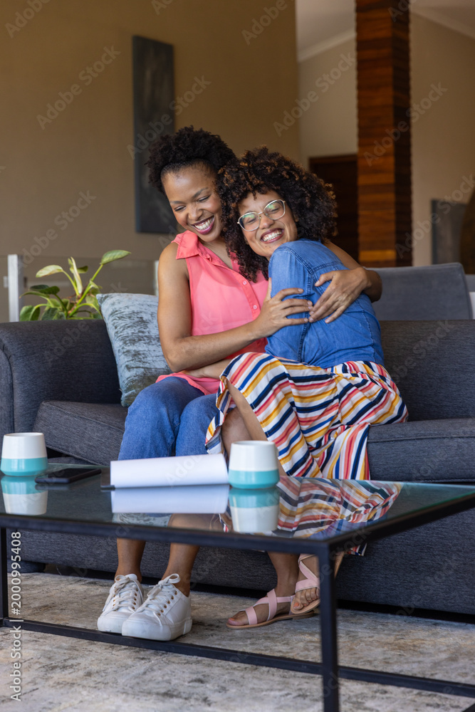 Fototapeta premium Diverse female friends sitting on gray sofa in living room, hugging, laughing with mugs