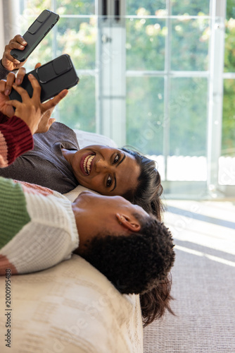 Diverse couple wearing knitwear lying on bed at home holding smartphones overhead, smiling together