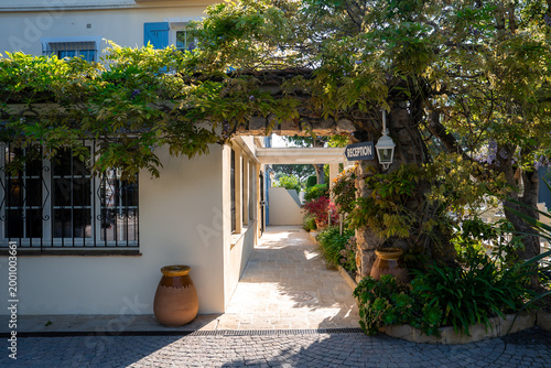 A sunlit garden path leads to a small reception under wisteria at a cream villa with blue shutters in Antibes, France. Terracotta urns and cobblestones frame the scene.
