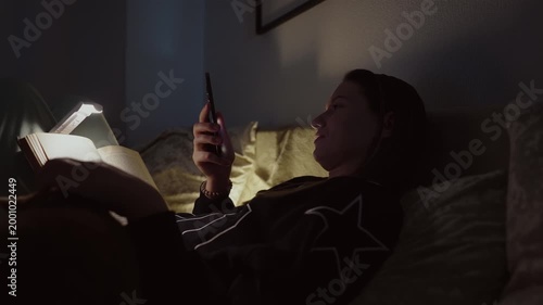 Girl doom scrolling her smartphone instead of reading on a coach at night. Social network and phone addiction concept. Cozy home ambient light