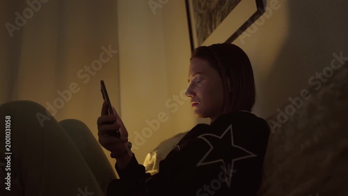 Girl doom scrolling her smartphone on a coach at night. Social network and phone addiction concept. Cozy home ambient light