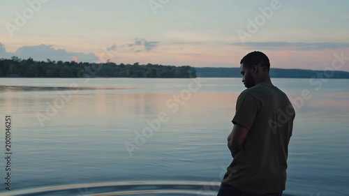 Adult black man standing near calm water, observing the serene landscape as ripples form in the water, reflecting the soft colors of the sky at dusk