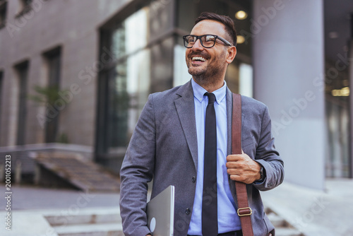 Schilderij op canvas Smiling Businessman In Suit With Laptop And Shoulder Bag Walking Confidently In