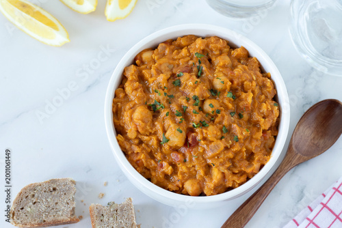 Принти на полотні Chickpea lentil curry in a bowl with wooden spoon, bread, and glass of water on white kitchen table