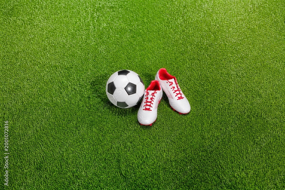 Naklejka premium Soccer ball and white athletic shoes with red accents placed on green artificial turf, showcasing sports equipment in a vibrant outdoor setting