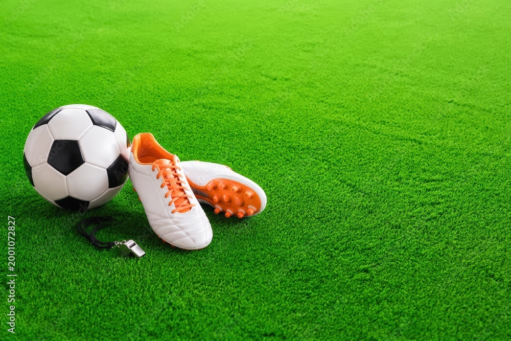 Naklejka premium Soccer ball and white cleats with orange accents placed on vibrant green artificial turf, whistle lying beside the shoes in a sports setting