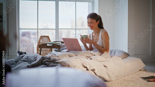 Lady having video call in bed drinking morning coffee. Happy brunette talking