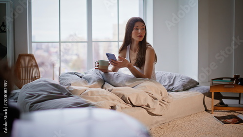 Awake girl scrolling smartphone in bed early morning. Relaxed woman sitting