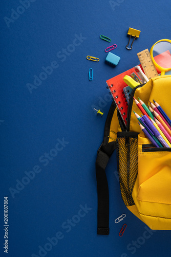 Yellow vertical backpack with colorful pencils, red notebook and school supplies on blue background for back to school