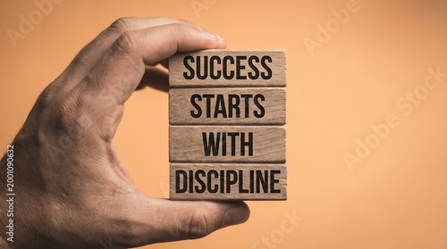 Hand holding stacked wooden blocks printed with success starts with discipline against a plain beige background presents a literal business motivation concept with copy space.