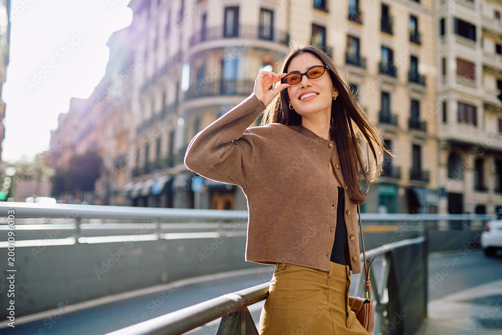 Fototapeta premium Young woman smiling, standing on a city street, lifting her sunglasses, enjoying the urban outdoor lifestyle and sunny day