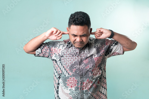 Schilderij op canvas An Asian man wearing a batik shirt covering his ears with fingers, expressing annoyance or avoiding loud noise and stressful information