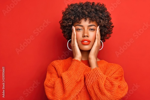 Confident young woman in orange sweater posing against a red background with bold makeup