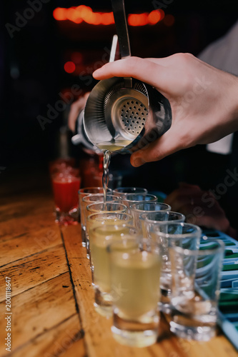 Bartender pouring fresh cocktails from shaker through strainer into multiple shot glasses on bar counter, close-up nightlife scene with warm lights, party drinks preparation concept