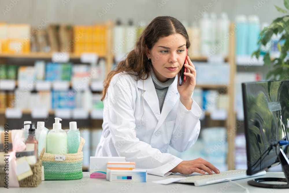 Fototapeta premium Young female pharmacist working with computer and talking on mobile phone in pharmacy