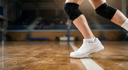 Close-up of volleyball player legs with knee pads and white shoes moving fast on a wooden indoor court during match.