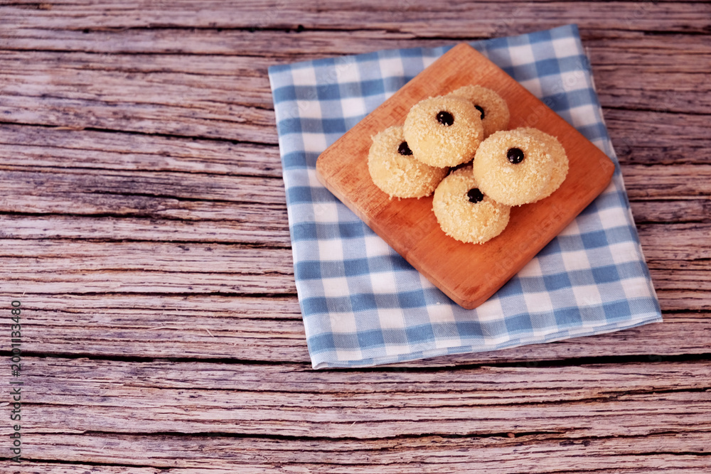 Fototapeta premium Kue Monde Susu or cookies rest on a wooden board. Board sits atop a blue-and-white checkered napkin. Napkin lies on a rustic wooden table. Ideal for cozy cafe or home baking themes