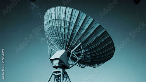 A dramatic photograph of a large satellite dish antenna on a tripod stand outdoors