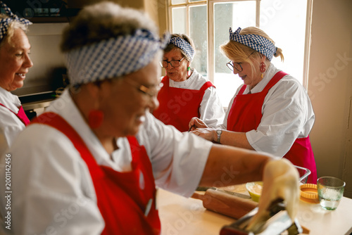 Elderly women of varied ethnicities collaborating on pasta making, showcasing ageless vitality and joyful engagement, illuminated by natural soft light.