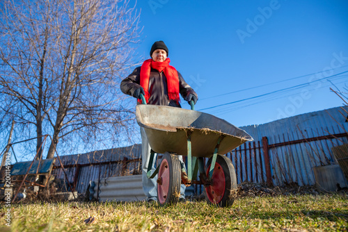 Man driving wheelbarrow in backyard garden. Gardener pushing cart for spring cleanup. Outdoor farm work, agriculture maintenance, seasonal yard task and landscaping maintenance concept.