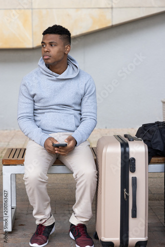 African American man sitting on bench in plaza wearing gray hoodie holding smartphone with suitcase