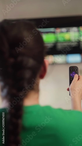 Young woman with braided hair holding a remote control, searching for content on a smart tv streaming service and choosing something to watch in a living room