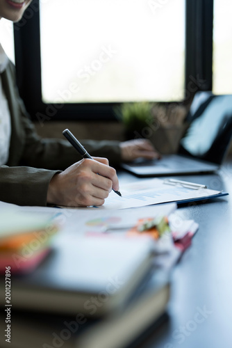 Close-up of businesswoman hand signing a contract or writing on a document while working at an office desk with a laptop.