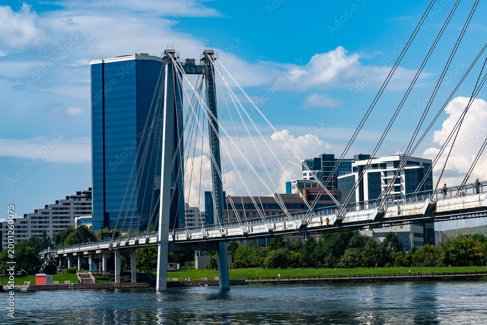Fototapeta premium Modern Cable-Stayed Pedestrian Bridge Over River With City Skyline and Glass Office Tower