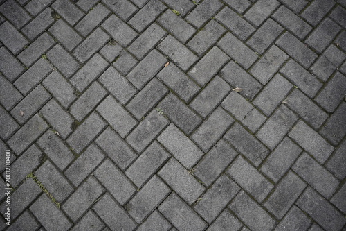 Top view of gray rectangular paving blocks in a herringbone pattern with small green weeds growing in the gaps. Urban pavement texture
