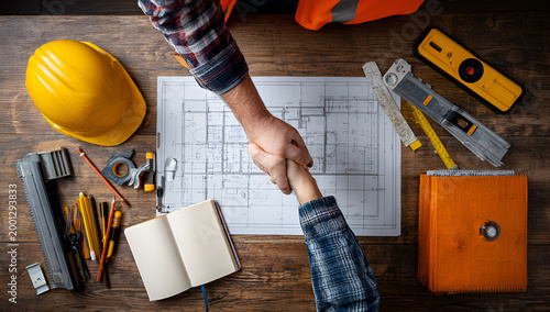 Construction Workers Handshake Over Blueprint and Tools on Wooden Table.