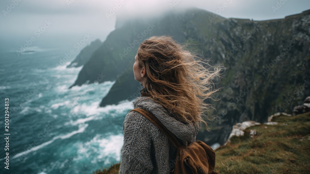 Fototapeta premium Woman with flowing hair gazes at misty cliffs and turbulent ocean waves under a cloudy sky, embodying a sense of adventure and solitude in nature