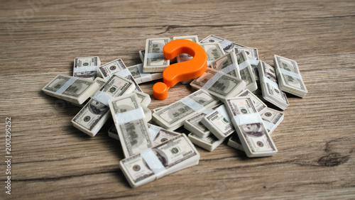 Orange question mark symbol sitting on a pile of hundred dollar bill cash bundles on a wooden desk.
