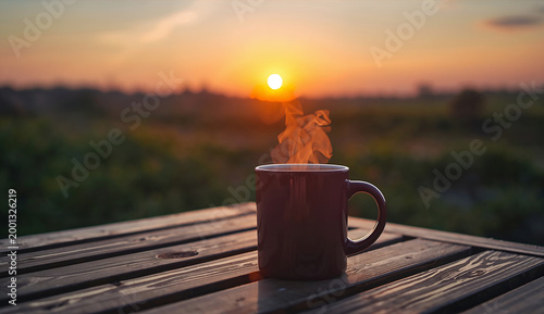 A steaming mug sits on a wooden table at sunrise, with a vibrant orange sky and silhouette of distant trees, evoking a peaceful, serene morning.