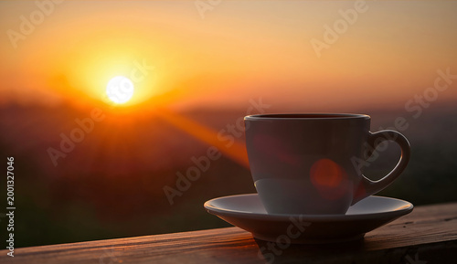 A white coffee cup on a saucer sits on a wooden surface, backlit by a vibrant sunrise. The warm glow creates a serene, peaceful atmosphere.