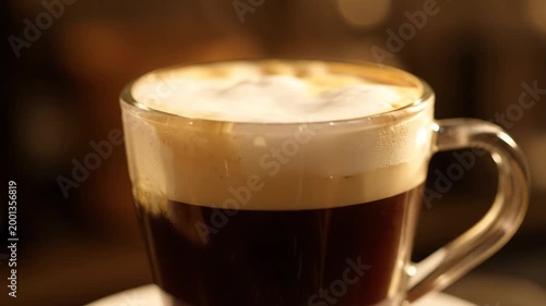 Close-up of layered espresso coffee drink with rich milk foam or cream top, served in a transparent glass mug under warm cafe lighting.