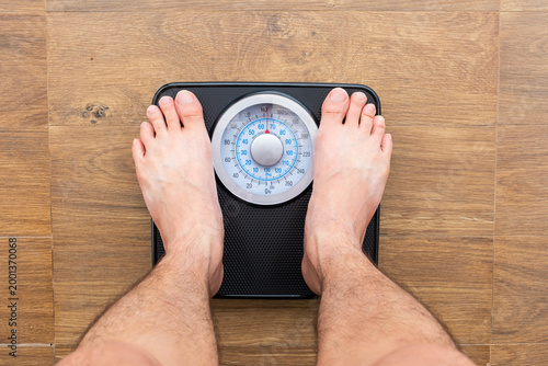 Bare feet of man standing on a black, analog bathroom scale on a light wooden floor. The person is checking their weight, concept focusing on health, fitness, and diet monitoring	
