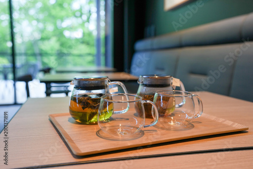 Glass teapot and cups rest on a wooden tray. Tea steams gently, green leaves swirl inside. Soft natural light streams through large windows. Modern cafe interior, calm and inviting