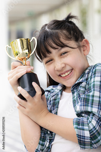 Confident, happy smiling young Asian grade-schooler or primary school girl holding a shiny gold trophy cup,  concept image for learning success, academic achievement, contest winning prize
