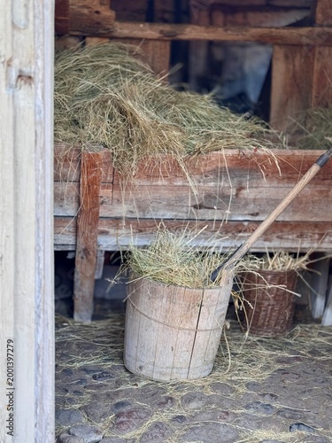 A rustic barn interior featuring a wooden bucket filled with hay and a hay fork leaning against it. The scene captures traditional farming life with natural textures and a calm rural atmosphere.