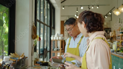 Senior Asian man and young woman in yellow aprons look at a tablet, managing stock in a sunny lifestyle cafe or craft shop. A small business team collaborating on orders and retail inventory.
