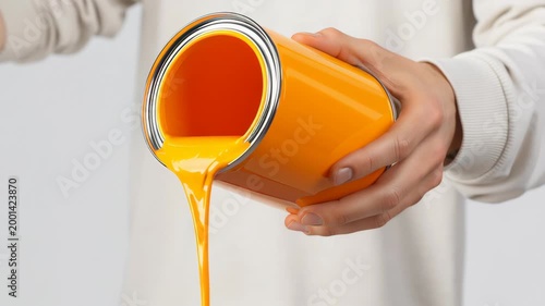 Person pouring bright orange paint from a can, showcasing the fluid motion of paint flowing out, with a neutral background emphasizing the action and color contrast