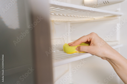 Housewife cleaning empty refrigerator shelf, wiping down interior with yellow sponge, focusing on hygiene, domestic chores and home kitchen sanitation. Concept of household chores and housework.