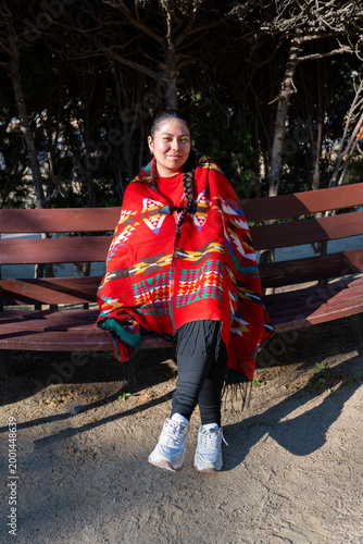 Peruvian woman sitting outdoors, wearing a red traditional poncho with indigenous patterns and looking at camera