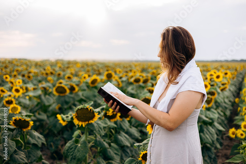 Woman reading Holy Bible in sunflower field at sunset