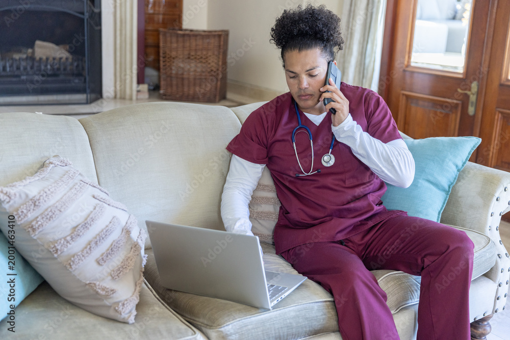 Fototapeta premium Mid-adult man in maroon scrubs sitting on sofa speaking on phone and using laptop with stethoscope