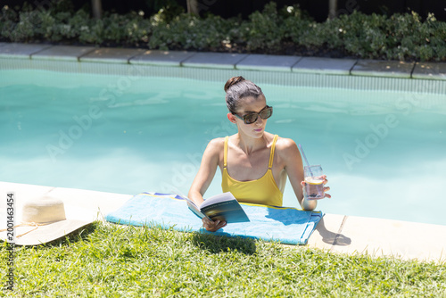 Mid adult woman leaning at poolside reading book holding glass with straw wearing yellow swimsuit