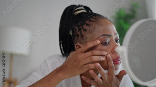 African American woman examines pimples on her face in a cozy room. 