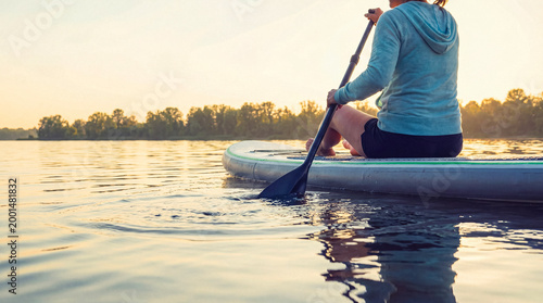Εκτύπωση καμβά Close-up photograph of a woman sitting on a paddleboard, gently paddling on a calm lake at sunrise or sunset